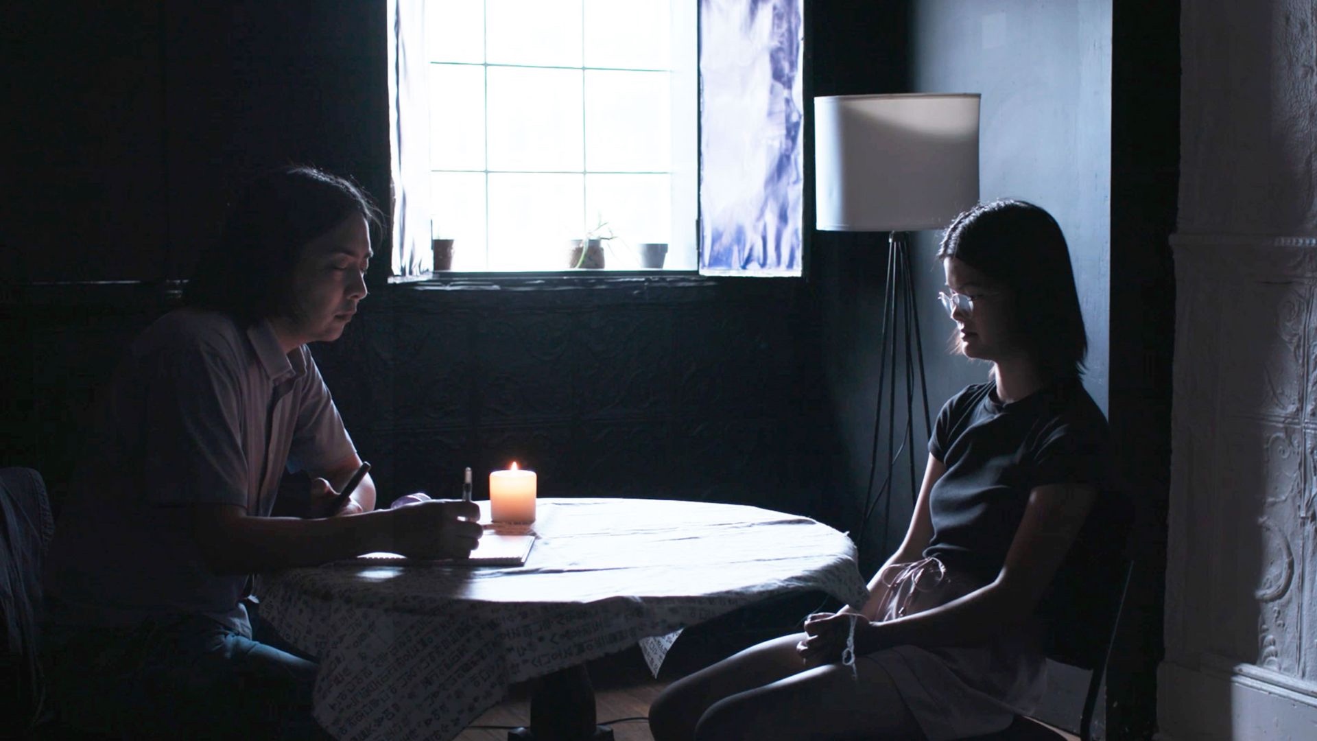 An Asian male writes in a notebook while seated across a table from an Asian female wearing glasses with her eyes closed.