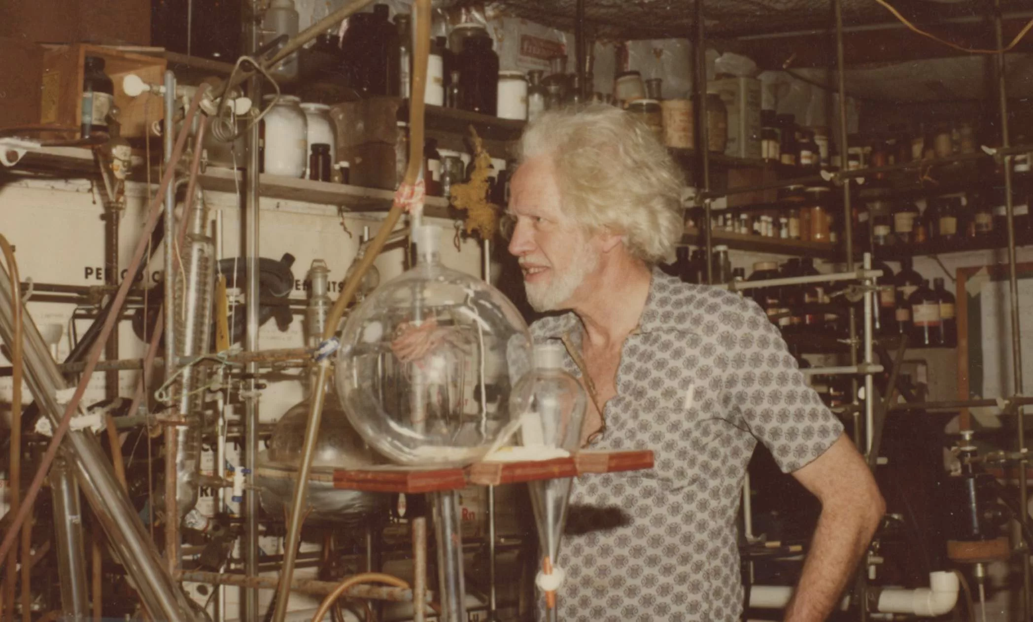 a man with a grey beard, grey hair, and wild look in his eyes stands in what appears to be a home-made lab.