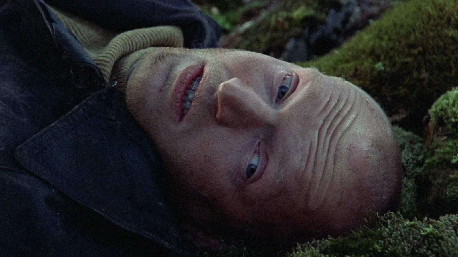 A man laying on the ground with little hair, wearing a blue jacket & green shirt stares blankly ahead.