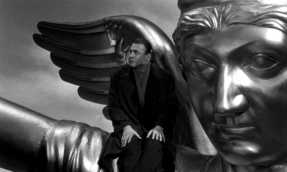 Iconic Bruno Ganz in a coat as a city's angel sitting by the wing of a bronze statue