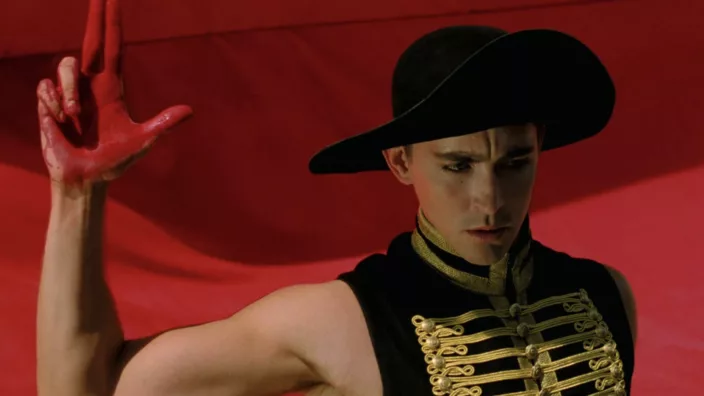 A man in a black, wide-brimmed hat & a black, gold-laced, sleeveless shirt with is right hand raised, smeared with red, in the shape of a gun, in front of a red background.
