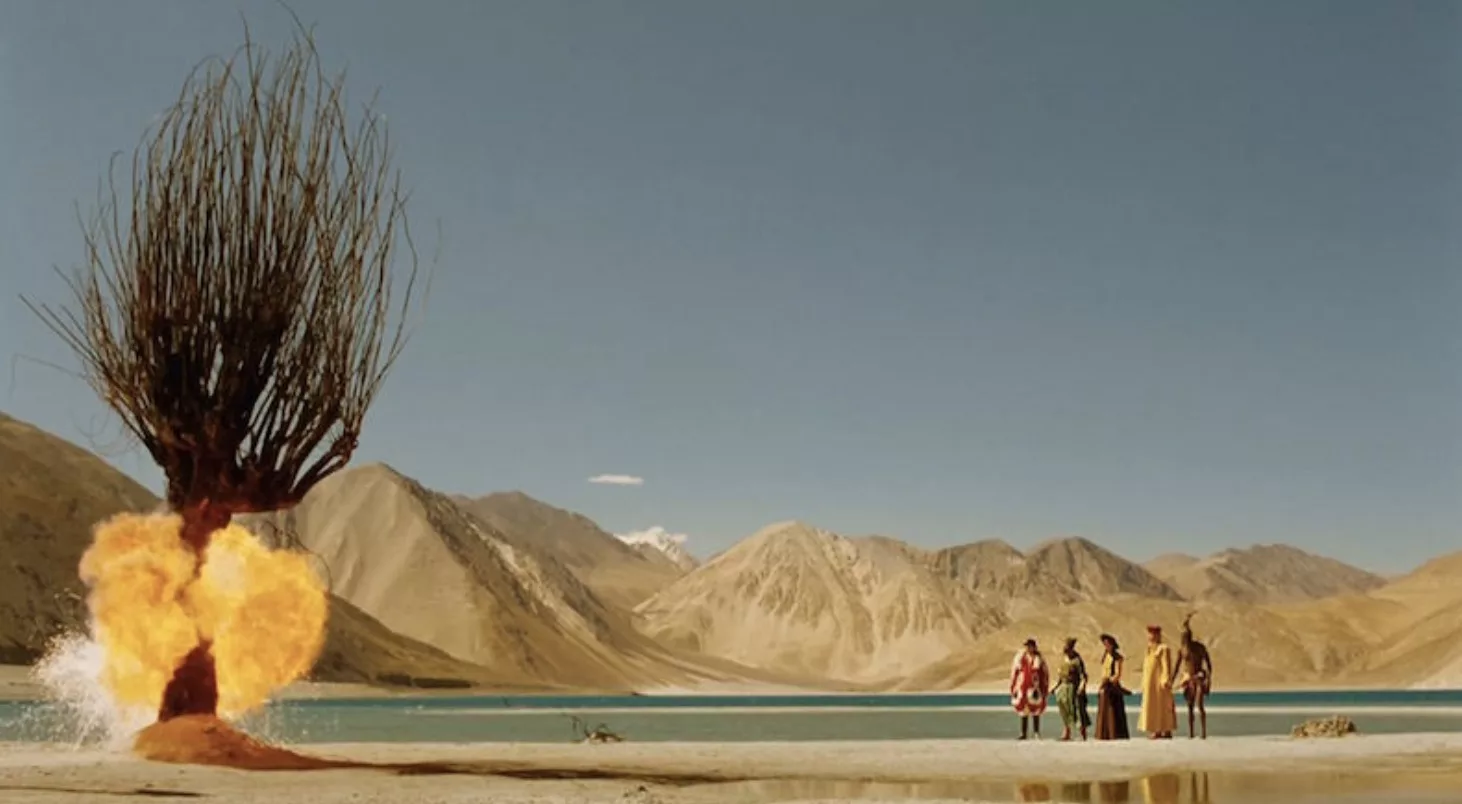 Five elaborately costumed people dwarfed by the surrounding mountains & desert, by a lake and what appears to be an exploding tree!