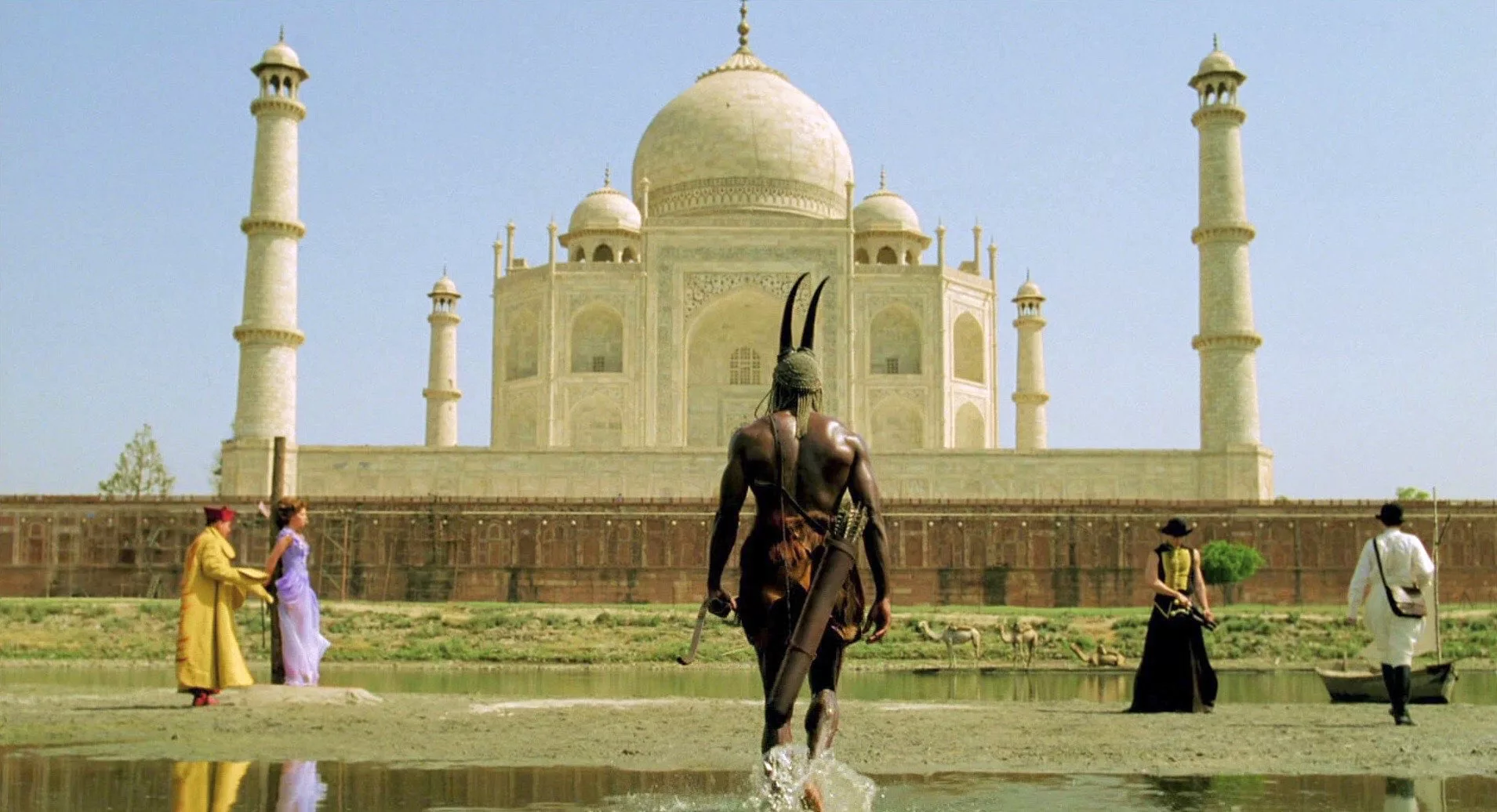 A large, dark man where a helmet with horns & a quiver of arrows slung 'round his shoulder, stands with his back us in front of the Taj Mahal, with a woman being tied to a pole on his left, a man in a black, wide-brimmed hat & a black, gold-laced, sleeveless shirt who appears to be holding a pistol & another man dress in white robes and a black bowler hat, to his right. Way in the background, 3 saddled camels await by a mote.