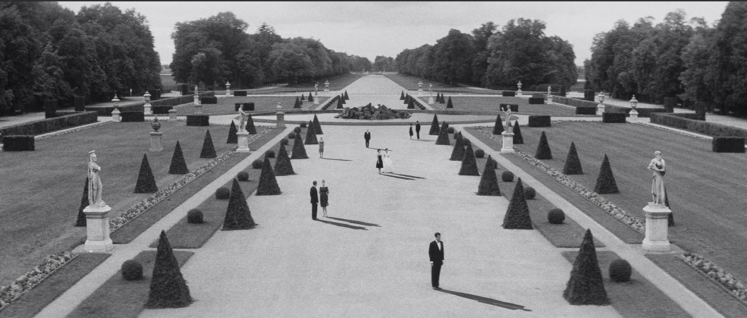 A black & white painting of a large, formal, elegant garden with shrubbery sculpted in the shape of pyramids, statutes, fountains, and lots of lawn. Interspersed throughout, men and women stand in dressed in formal wear, casting long shadows.