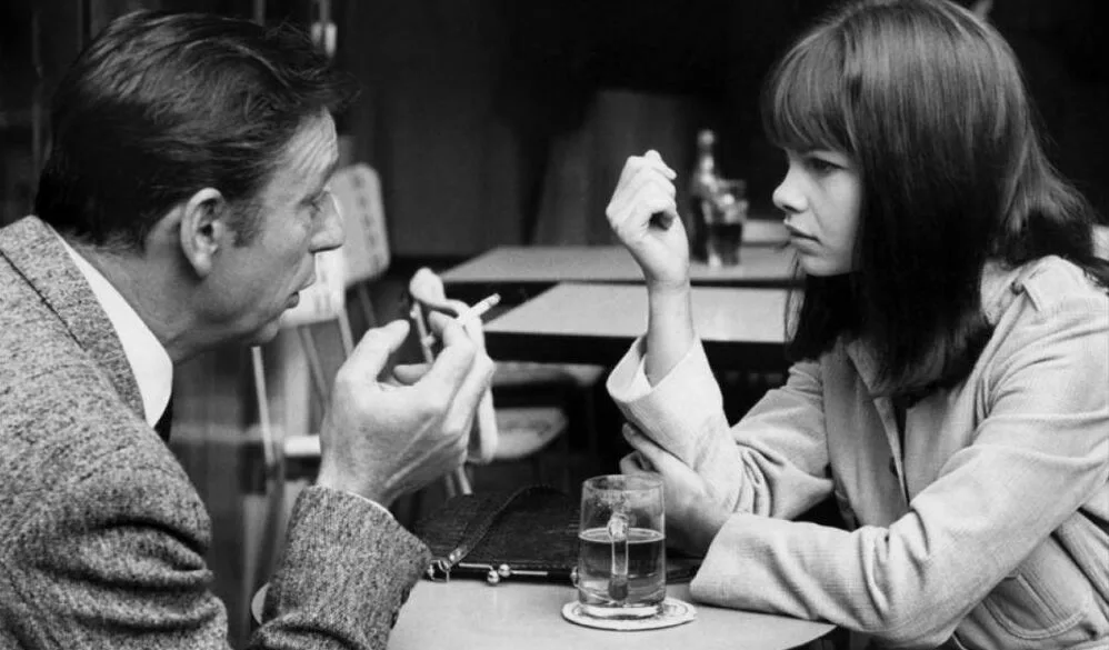 A black & white image of a middle-aged man in a bar, cigarette in right hand, looking intently at a young woman, with empty tables in the background. From the look in their eyes, they might be scheming revolution.