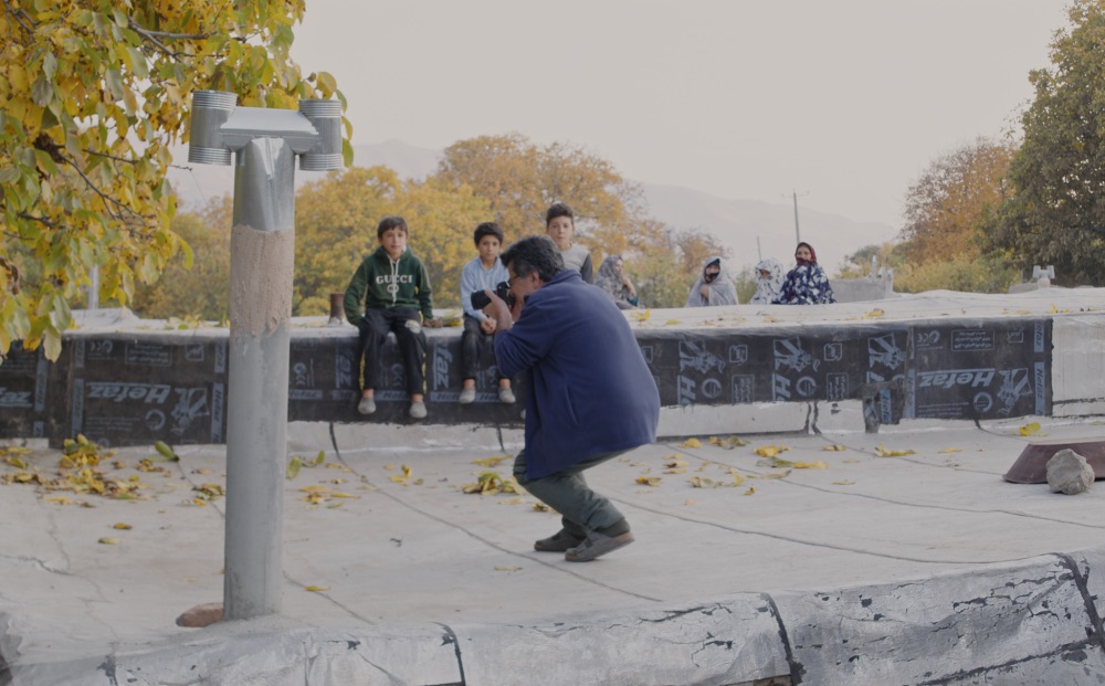A man crouches with a camera. Kids sit on a wall in the background