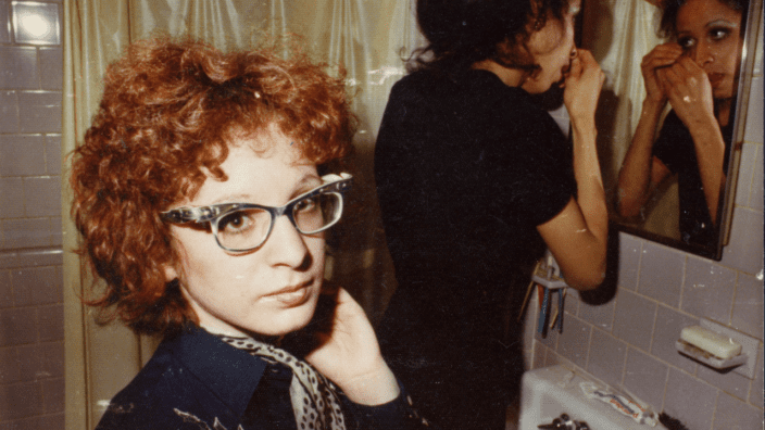 a woman with red head and glasses looks at the camera while another woman looks in a bathroom;s mirror behind her.
