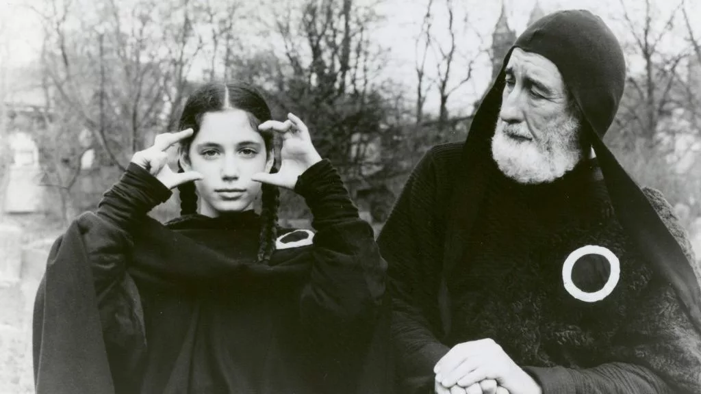 A young girl in pigtails and a black robe holds both hands near her face as an older, bearded man in black robes and cap looks toward her with an expression of concern