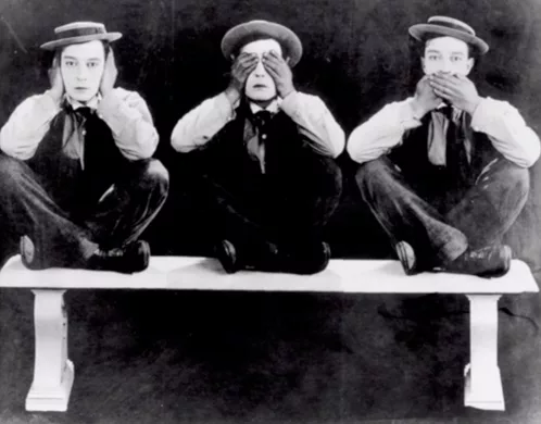 Three identical men sitting on a white bench and each either covering their ears or eyes or mouth with hands