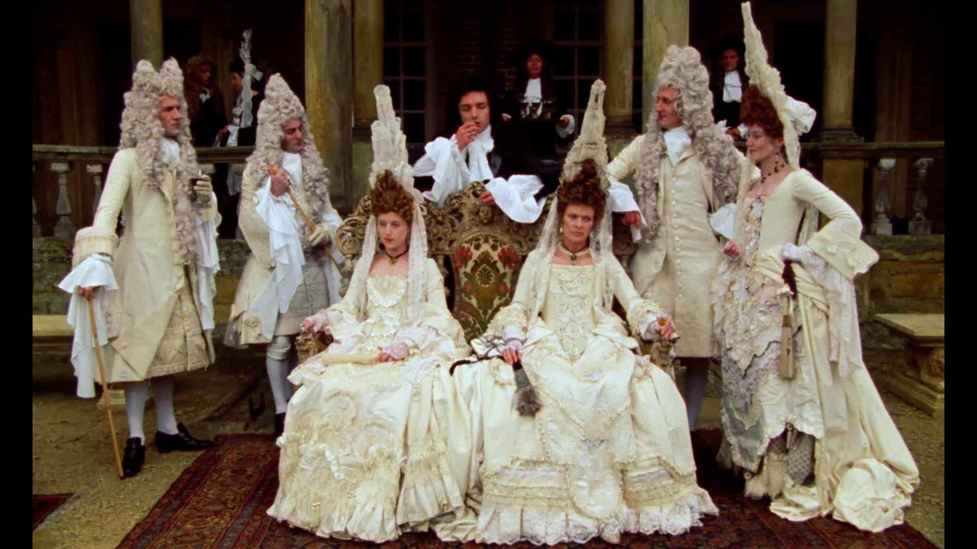 Two royally dressed aristocratic women settled in chairs as they are surrounded by their confidants