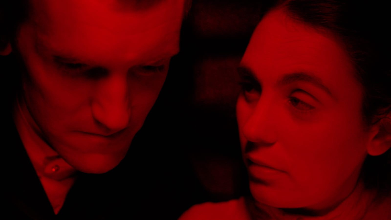A young man, looking down and a woman looking at the man, both bathed in red light