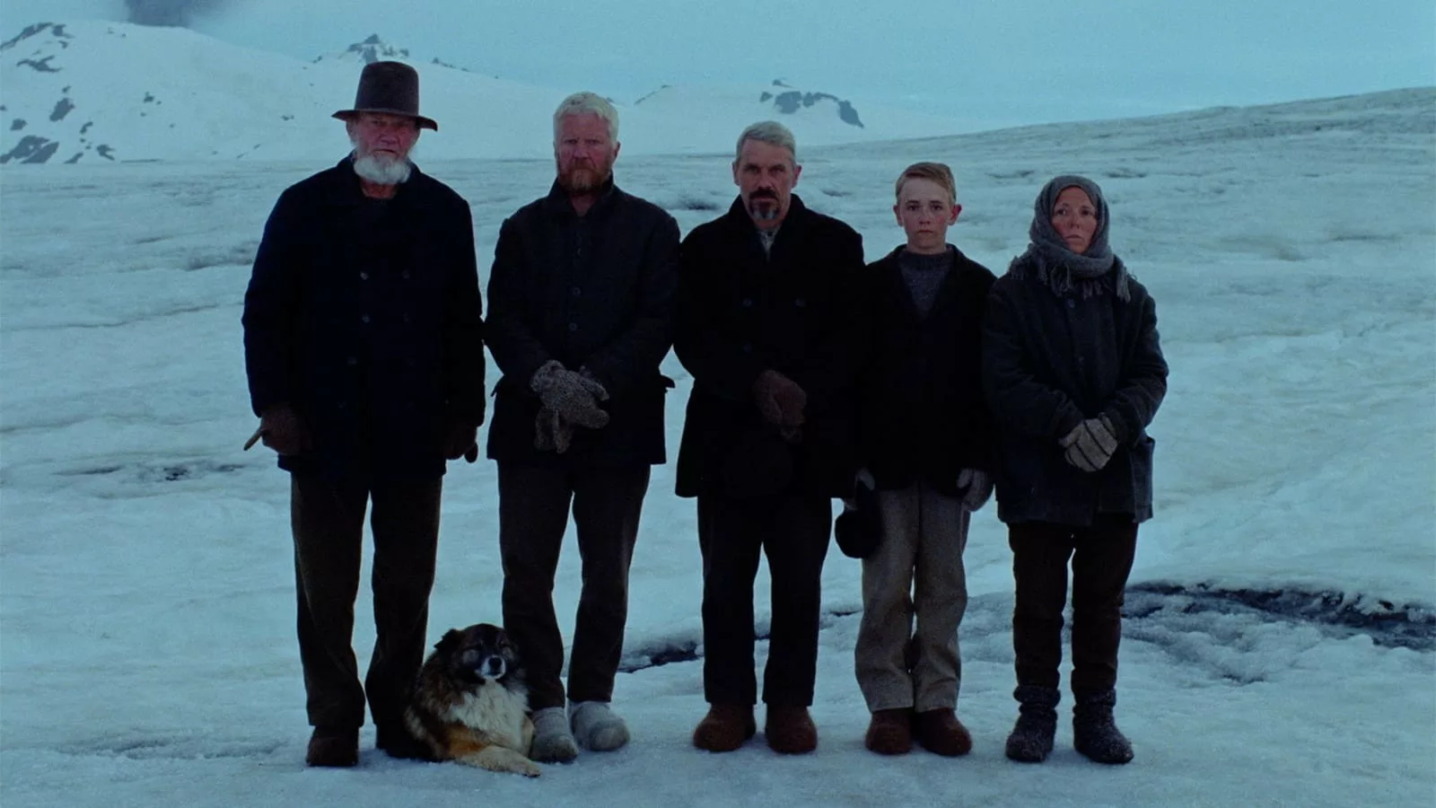 3 men, 1 child, 1 woman, all dressed in early 20th Century garb and a dog stand on a desolate ice field with mountains in the background