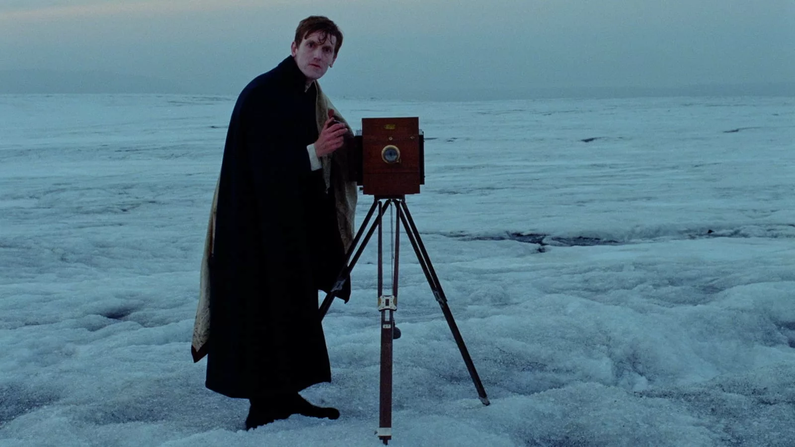 A young man in a cape and boots stands aside an old fashion camera, on a large, desolate ice field