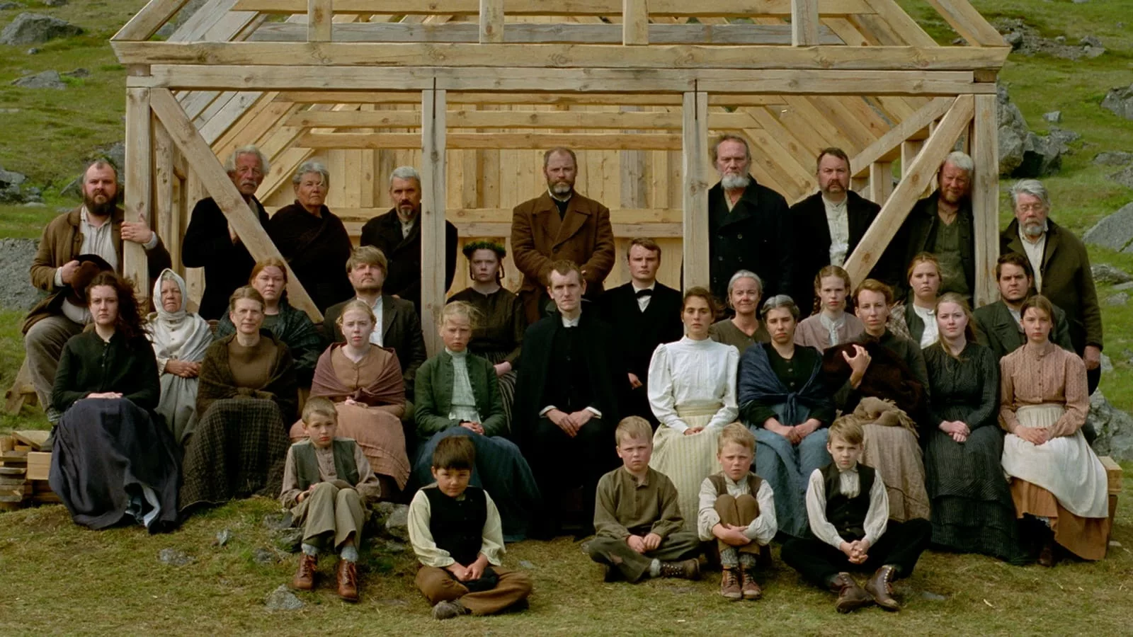 A large group of men, women & children, all dressed in early 20th century garb pose for a picture in front of the frame of a building under construction
