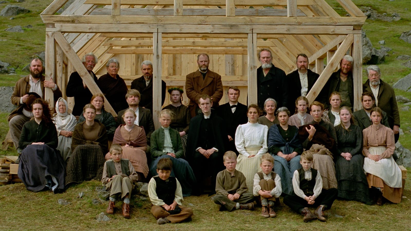 A large group of men, women & children, all dressed in early 20th Century garb are posed in front of the frame of a building under construction.