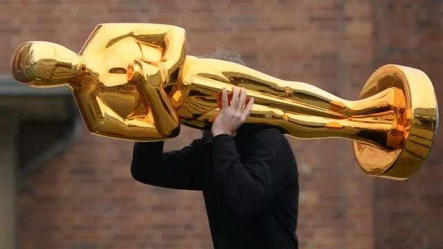 Someone dressed in black with their head obscured by the giant Oscar statue on their shoulder