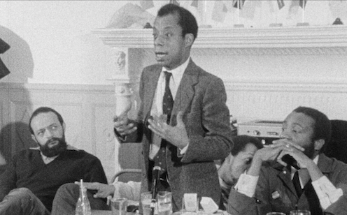 Two men listen to James Baldwin delivering a heartfelt speech in a suit