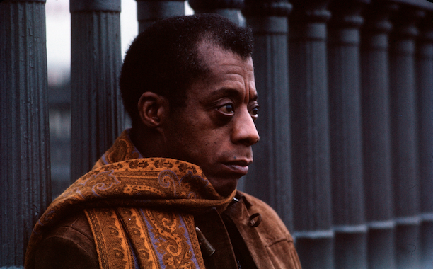 James Baldwin in an orange scarf staring in the distance from a columned bridge