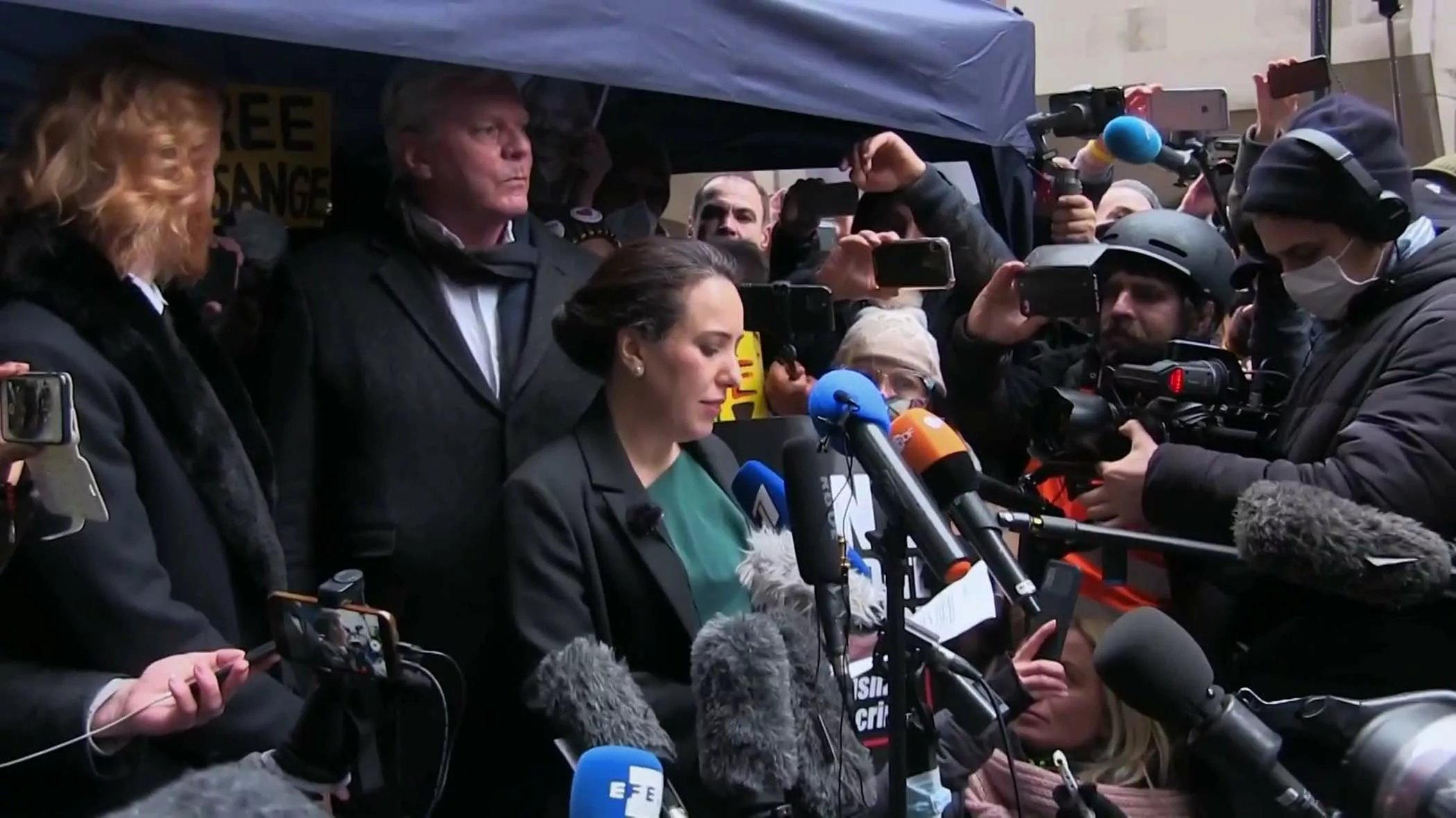 Hundreds of journalists and cameras attacking the spokespeople with microphones for comments on their exit from a tent with Free Julian Assange slogan