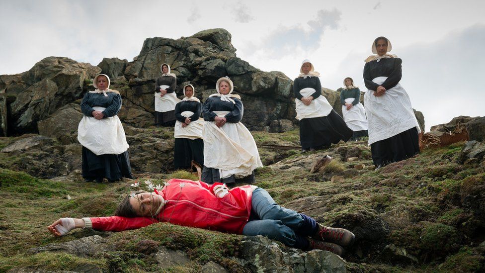 A middle-aged woman in a read wind jacket lying on the rocky soil with the camomiles in front of her, watched by a few nuns in white aprons behind her
