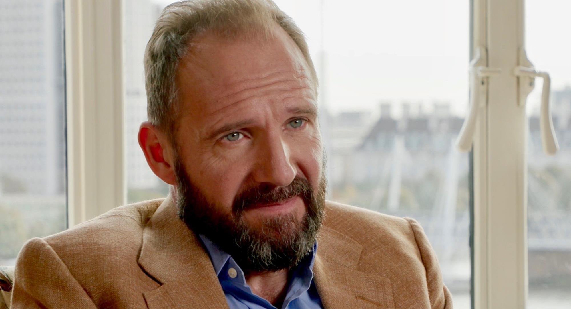 Bearded Ralph Fiennes in a blue shirt paired with a brown jacket against a window