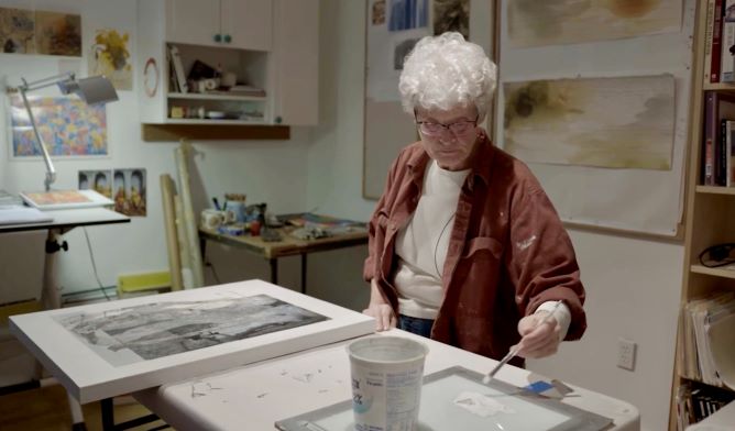An old grey-haired woman painting with a brush in a workshop full of inks, paints, and pictures of pastel-colored landscapes