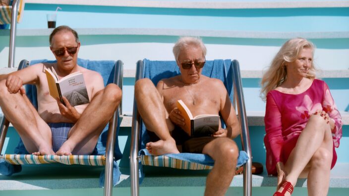 Two bare-chested balding men in speedos reading the same book as they enjoy their vacation with a gray-haired woman in a pink summer dress sitting closeby