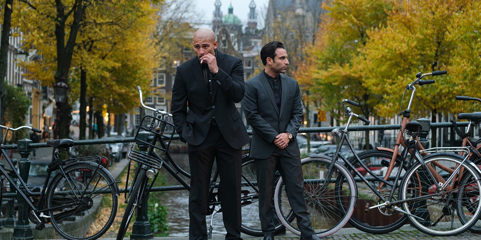 Two gangster-looking men in black suits on a small, though filled with bikes, bridge across the channel