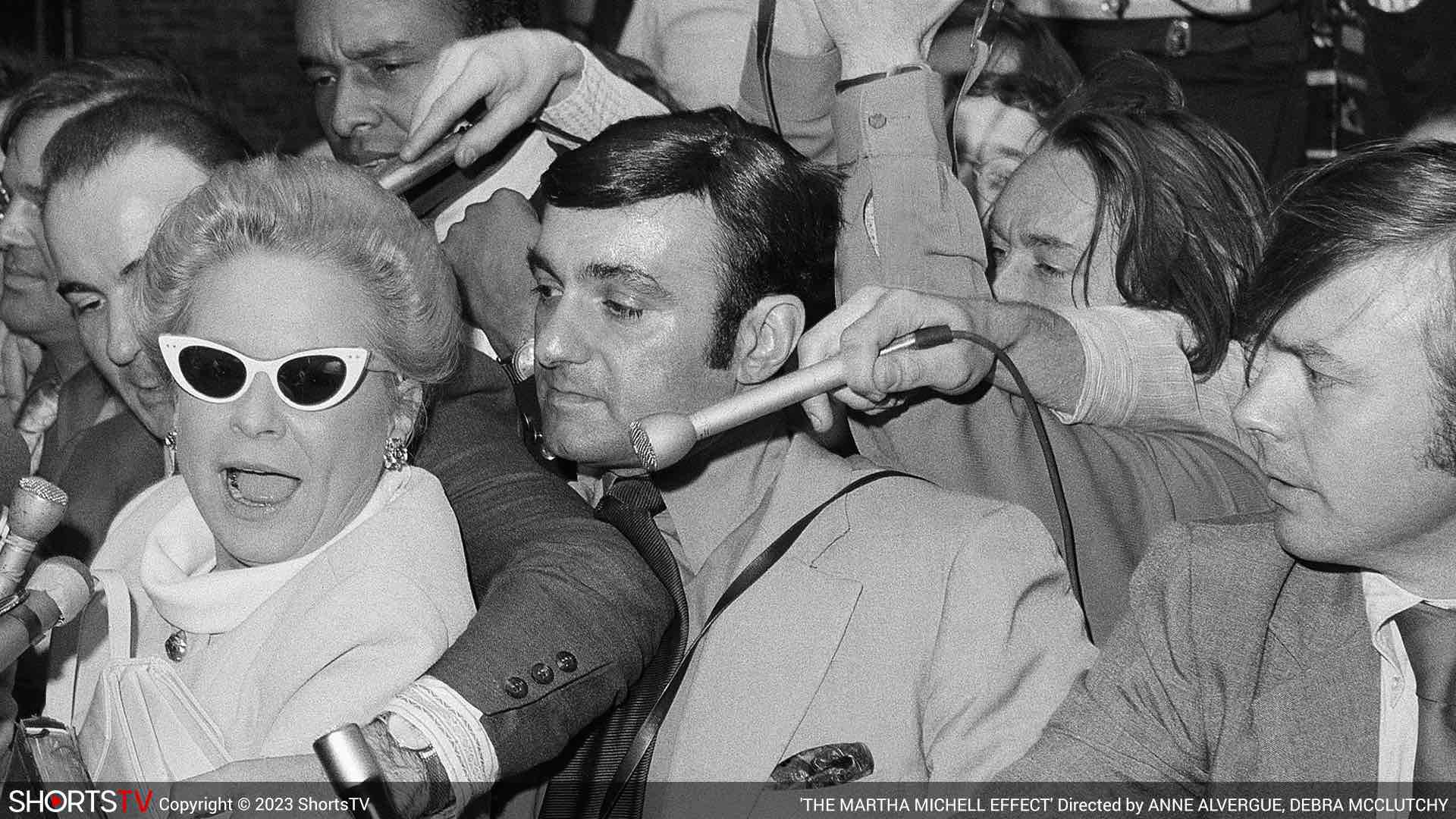 Martha Mitchell in black sunglasses attacked by multiple reporters as they point their microphones to interview her in The Martha Mitchell Effect, an Oscar 2023 nominated short documentary