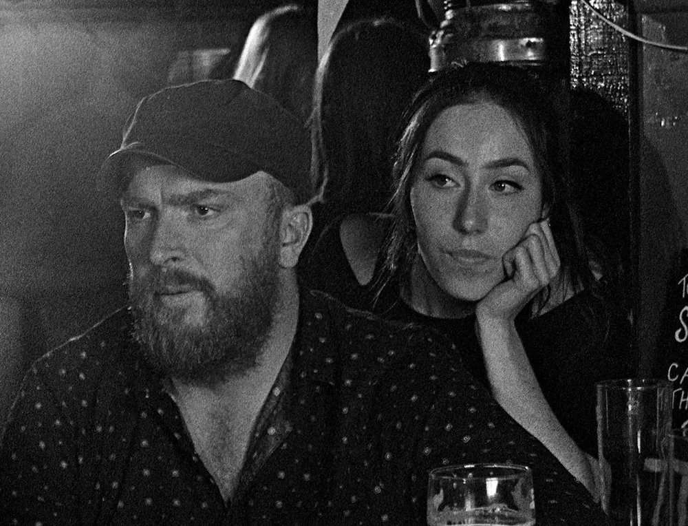 A bearded man in a black hat and a polkadot shirt speaking with someone as a young woman intimately behind him follows their conversation, her head resting on her arm