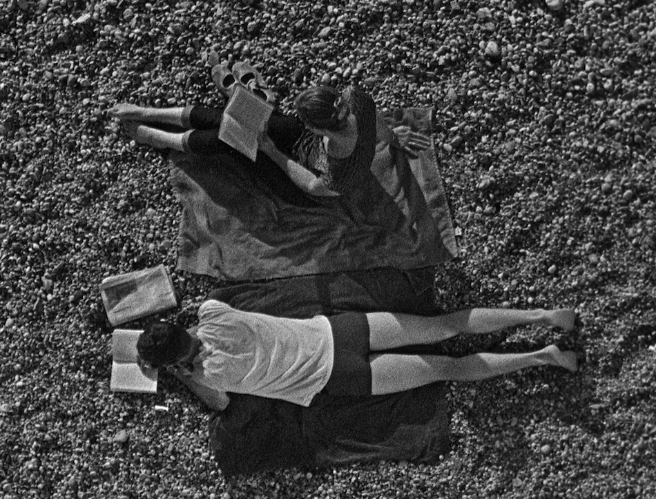 A man lying on a blanket in his underpants and a woman sitting beside him as both read their books on a pebble beach, shot from above