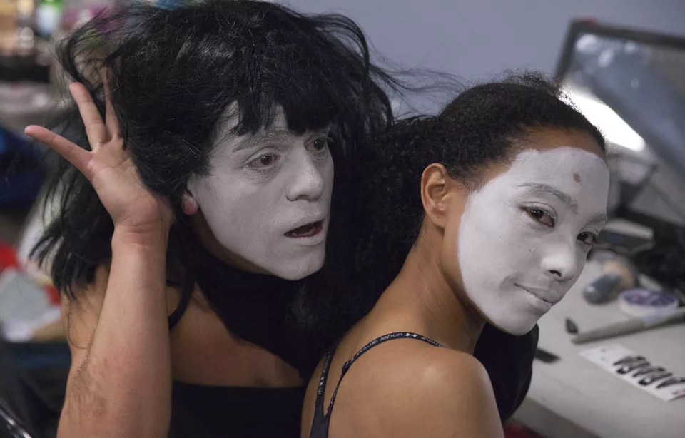 A man in a black wig and a woman, both with faces painted in white, at a makeup stand in a dressing room as they are getting ready for a show