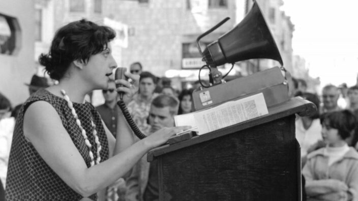 A woman in a sleeveless dress speaking on the speakerphone before she is to deliver a speech to the crowds of people around her stand