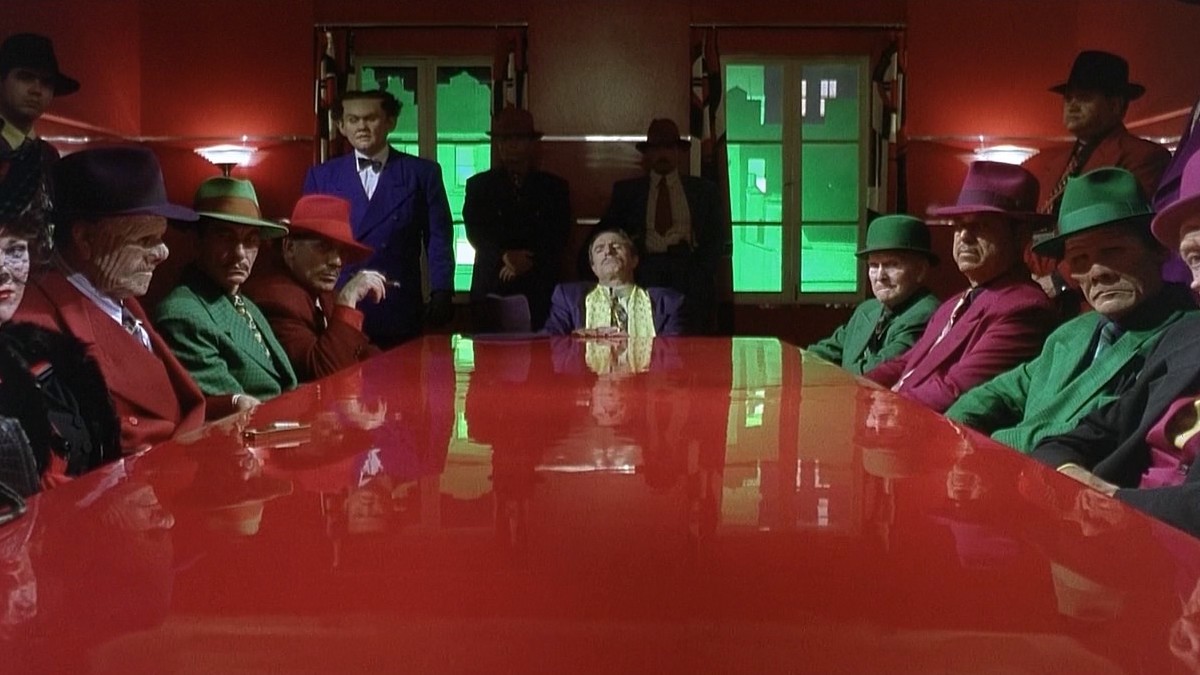 A group of ugly-faced men in bright overalls sitting around a big red table in a gloomy room lit with harsh green light pouring through the windows