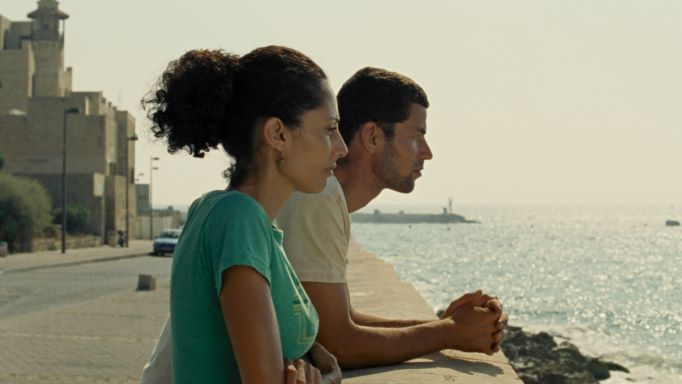 A man and woman with dark olive skin and brown hair look off into the distance. The sea is in the background.