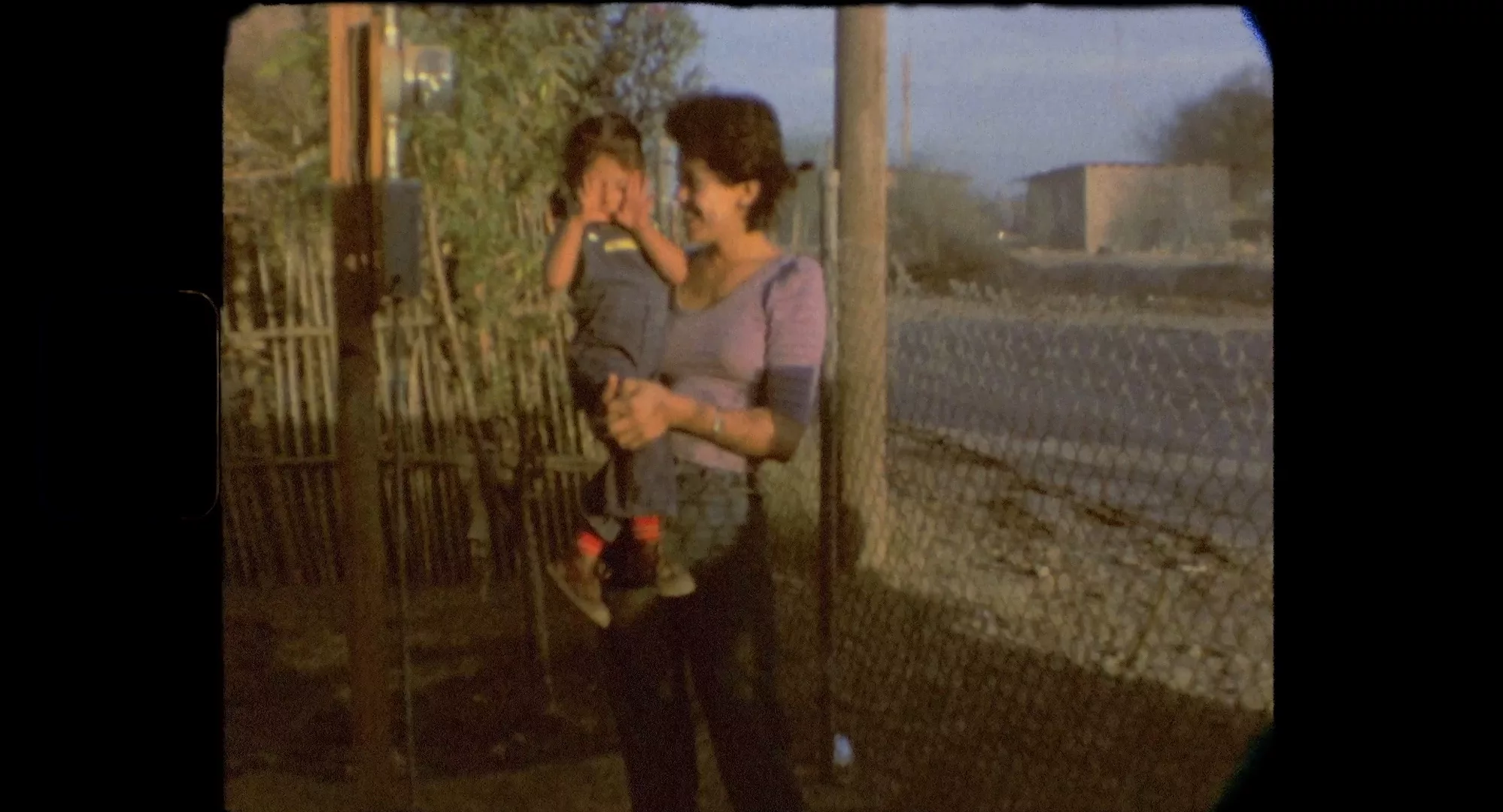 A old film still with a woman holding a child. The child hides her face in her hands. They are standing on a sidewalk and the sky is blue in the background.