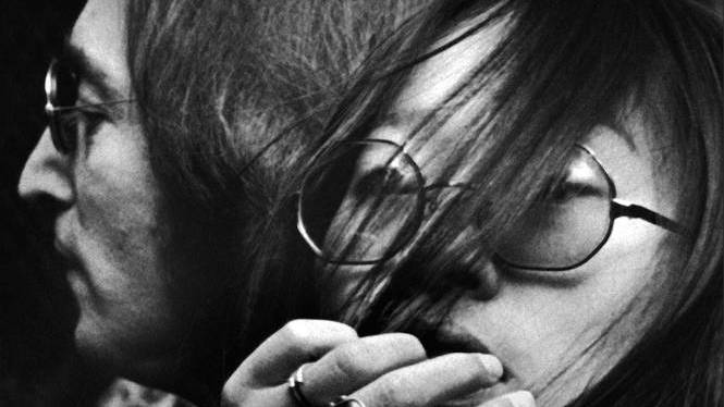 May Pang leaning towards John Lennon with her hand above her mouth as he looks away from the camera, both in spectacles