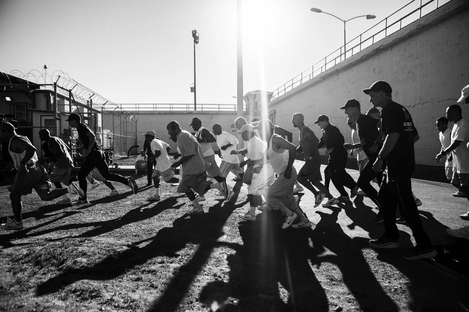 Black and white image of 20 men running with sun brightly shining in the background and long shadows. You can see the prison walls and barbed wire in the background.