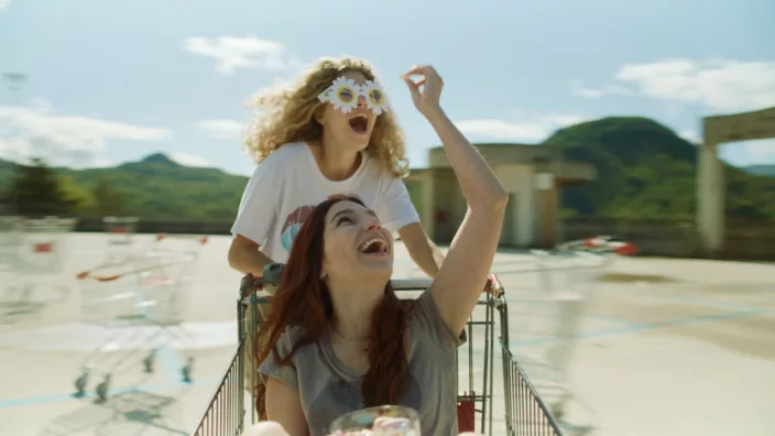 Blonde girl in camomile-framed glasses having fun with another girl as she sits in a shopping cart