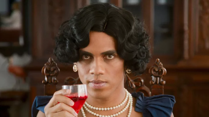 Trans woman with a pearl necklace and luxurious earrings raises a glass of red liquid as she looks in the camera