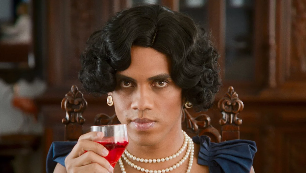 Trans woman with a pearl necklace and luxurious earrings raises a glass of red liquid as she looks in the camera