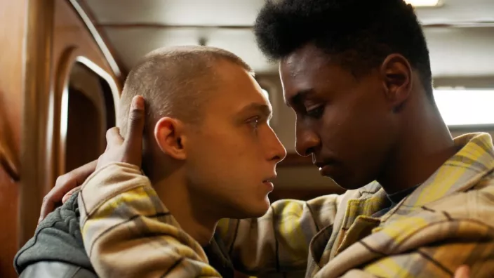 Black guy in a hoodie embraces a white guy as they are about to kiss while traveling on a train