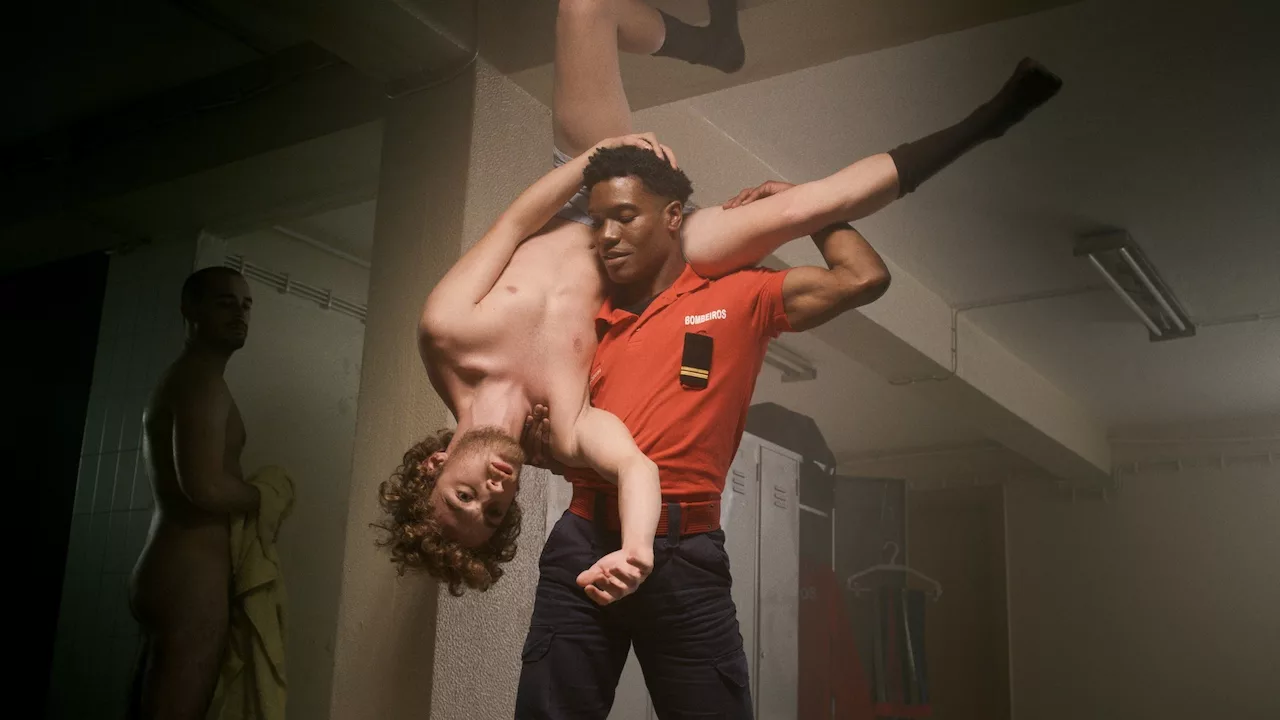 Black firefighter holds up a naked amateur guy in a figure as another firefighter watches them