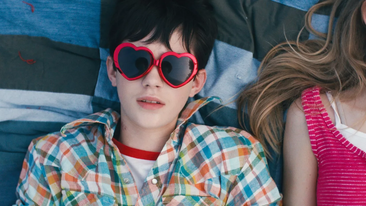 Black-haired boy in red-framed sunglasses looks in the camera as he lays on the grass with a girl