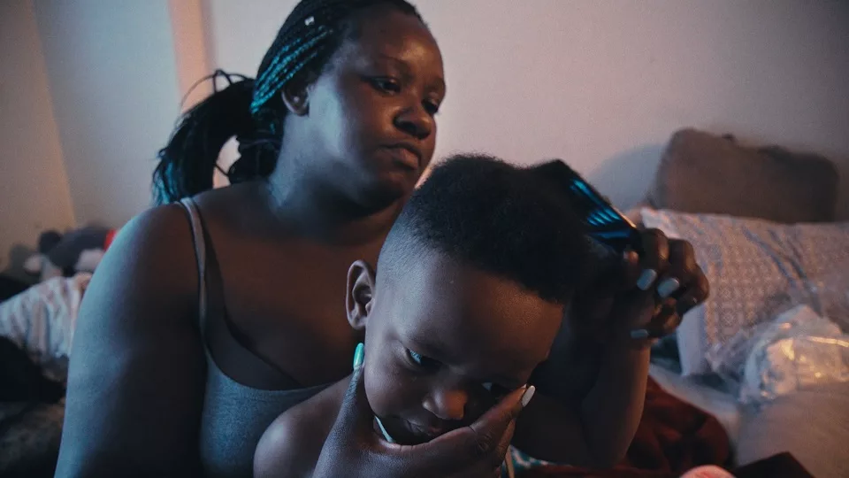 A Black woman and boy sitting on a bed. The woman combs the boys hair. The woman has bright blue fingernails.