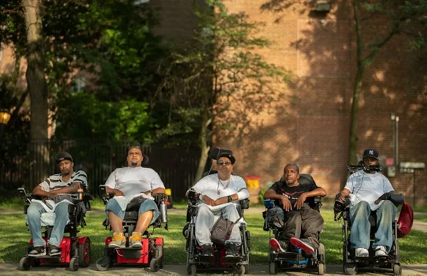 Group of five men in wheelchairs looking at the camera. They are on grass with a brick wall behind them.