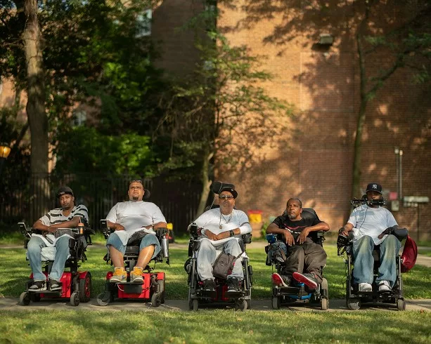 Group of five men in wheelchairs looking at the camera. They are on grass with a brick wall behind them.