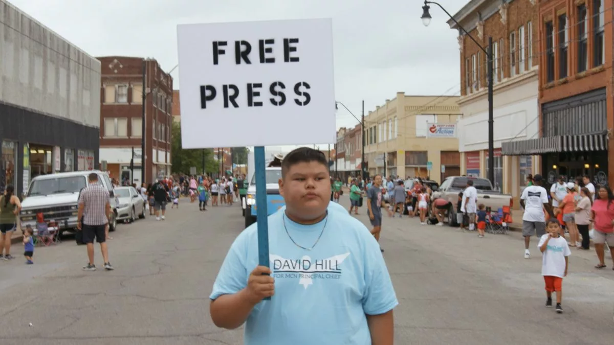 Young boy stands in street in light blue shirt with a picket sign that reads "Free Press"
