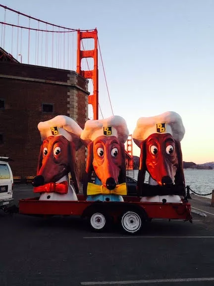 three oversized dog heads with big eyes and long ears and noses are on a trailer in front of the golden gate bridge.