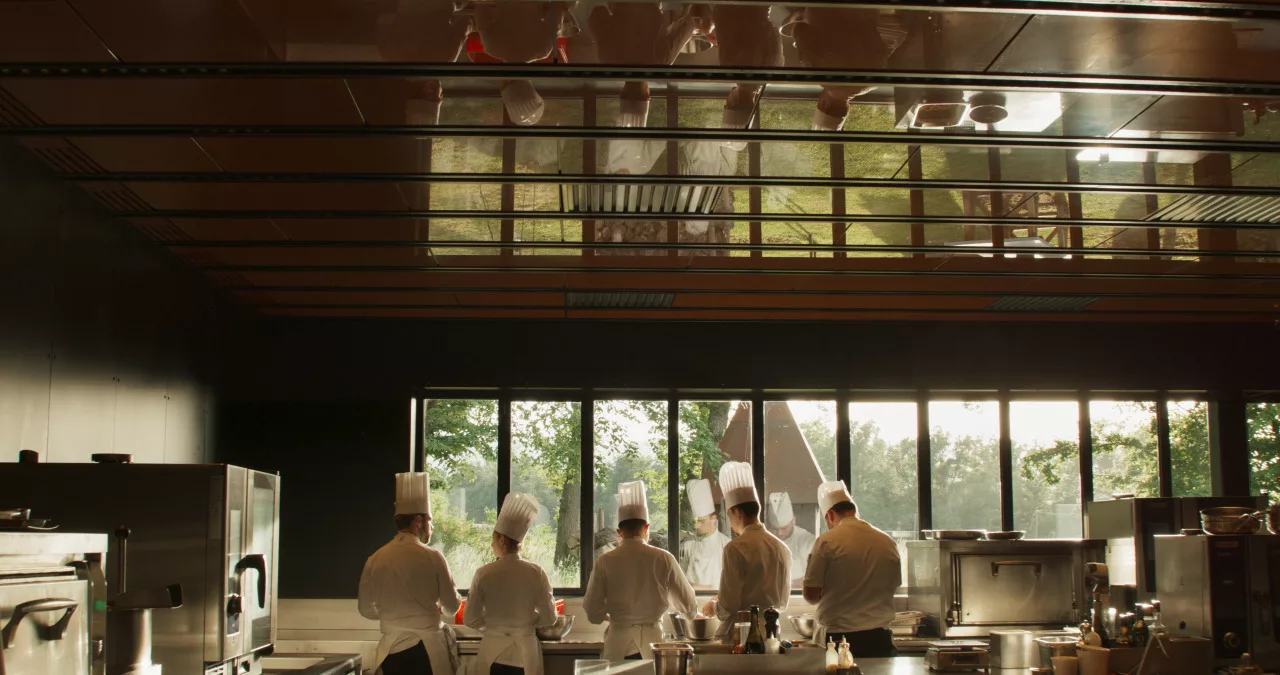 Five chefs, all dressed in whites and "Toque Blanche" standing side by side in a kitchen, facing a window reflecting their faces.